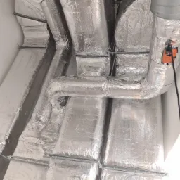 Izolowane, srebrną folią aluminiową, kanały wentylacyjne pod sufitem, z widocznymi elementami mocującymi i białym anemostatem.