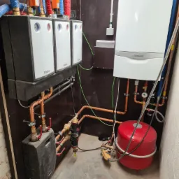 Zestaw urządzeń grzewczych Vaillant zamontowany na ciemnej płycie, widoczne trzy moduły zasilania, piec gazowy, czerwony zbiornik wyrównawczy, oraz liczne miedziane rury i zawory.