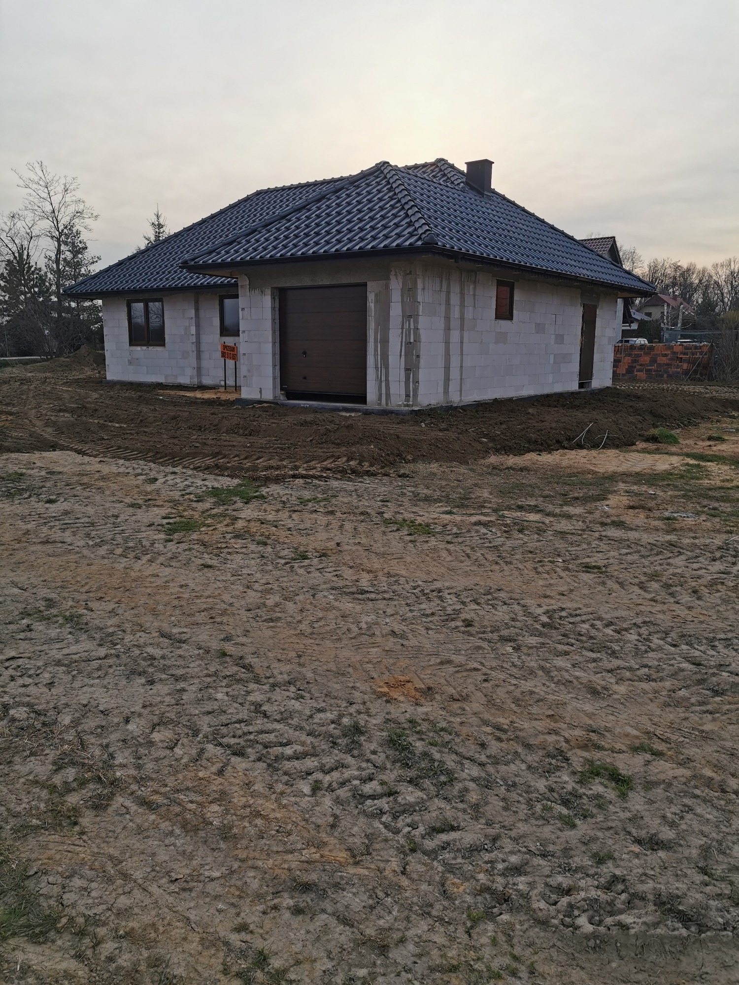 Surowy stan parterowego domu z bloczków, z ciemną dachówką, brązową bramą garażową i przygotowanym terenem wokół.