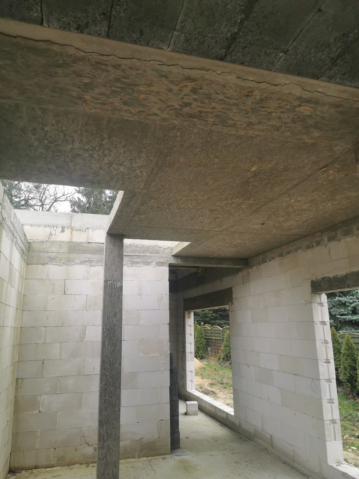Wnętrze surowego domu w trakcie budowy, widoczne ściany z betonu komórkowego, betonowe słupy i strop z płyt OSB, na zewnątrz widok na zieleń.