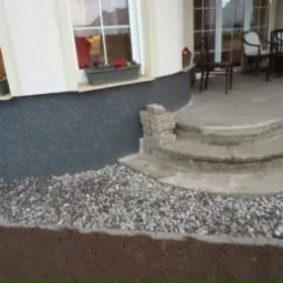 Wykończony fragment elewacji domu z oknem, schodami i tarasem. U podstawy elewacji ozdobny pas z kamieni oddzielony obrzeżem od ziemi.