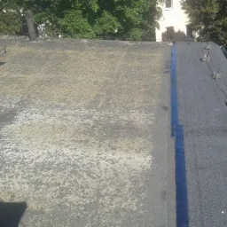 Widok z góry na płaski dach pokryty papą, z wyraźnym podziałem na starą, miejscami porośniętą mchem i nową, świeżo położoną część z widocznymi zabezpieczeniami odgromowymi.