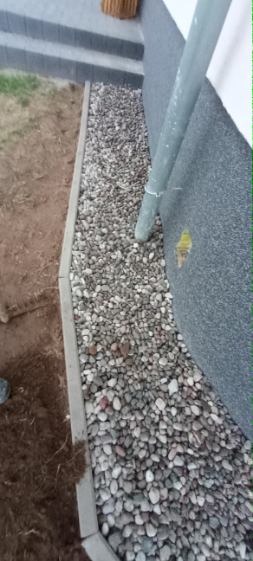 Wykończenie rabaty przy ścianie budynku drobnymi kamieniami, oddzielonej obrzeżem betonowym od ziemi. Rynna odprowadzająca wodę.