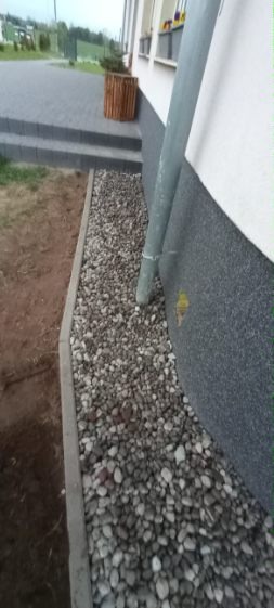 Drenaż z kamieni przy ścianie budynku, obrzeże z kostki, rynna spustowa. Donica z rośliną w tle. Widok na fragment chodnika z kostki brukowej i trawnik.