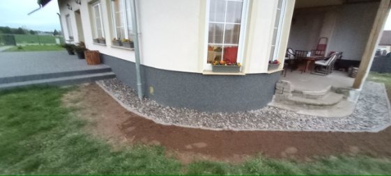 Obrzeże z kamyków oddziela trawnik od elewacji domu z oknami i schodami. Na parapecie okna skrzynka z kwiatami. Taras z meblami ogrodowymi.