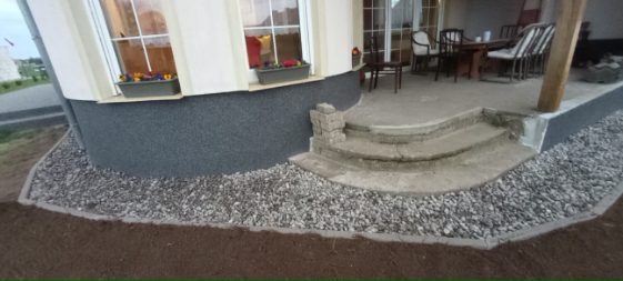 Wykończony fragment elewacji domu z oknem, schodami i tarasem. U podstawy elewacji ozdobny pas z kamieni oddzielony obrzeżem od ziemi.