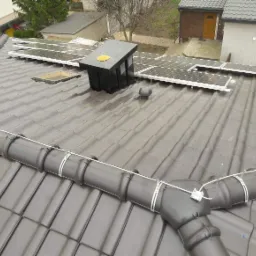 Szary dach pokryty dachówką ceramiczną z zainstalowanymi panelami słonecznymi, kominkiem wentylacyjnym i systemem rynnowym. Widoczne mocowania paneli i rur. W tle budynki mieszkalne.