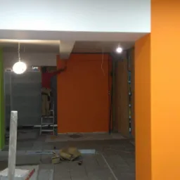 Pracownik na drabinie maluje pomarańczową ścianę w nowo budowanym lub remontowanym wnętrzu, widoczne niedokończone podłogi i surowe wykończenie.
