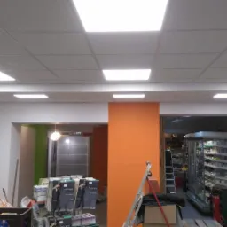 Sufit podwieszany z kwadratowymi panelami LED w sklepie w trakcie remontu, widoczne materiały budowlane i drabina.