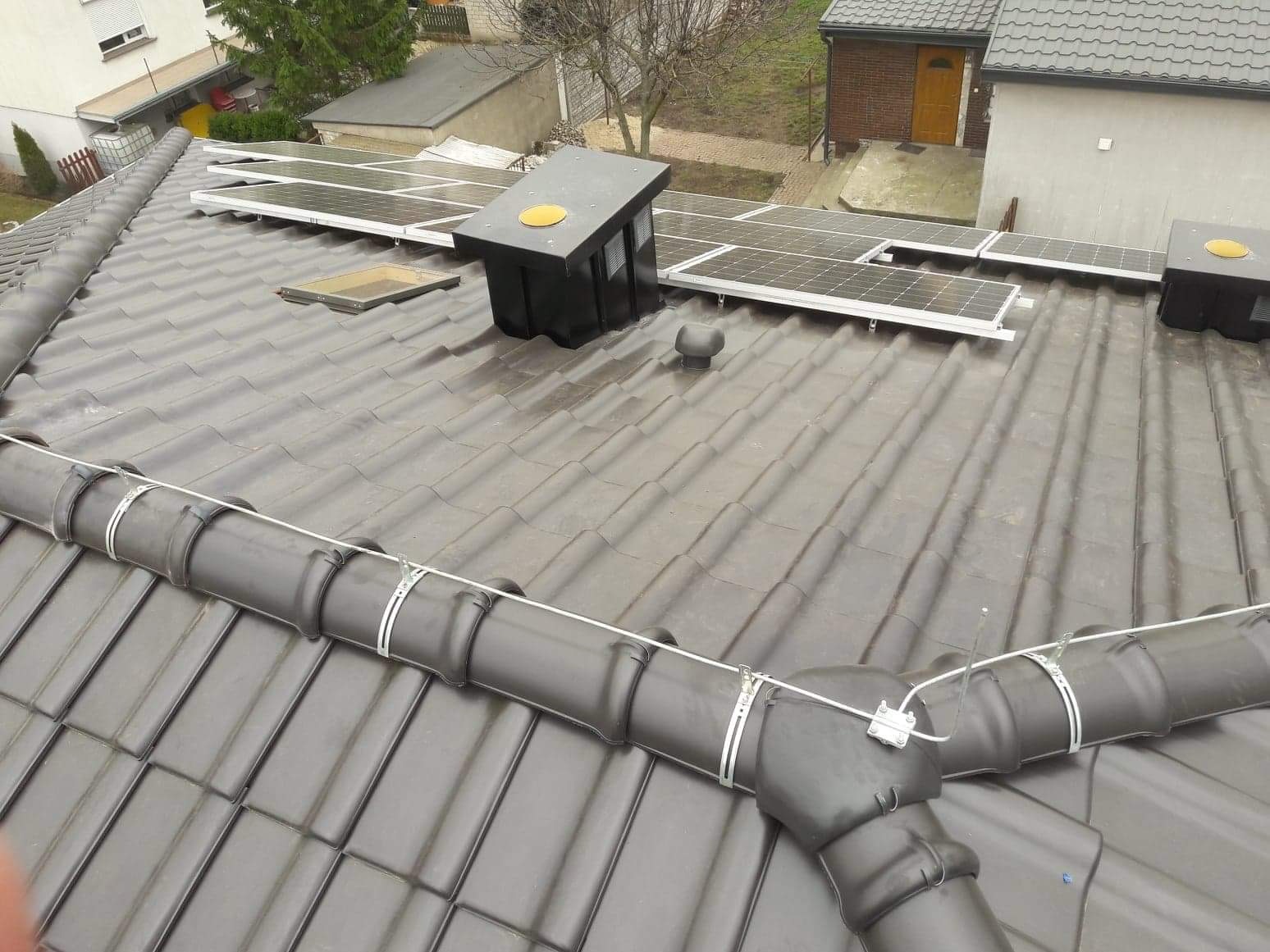 Szary dach pokryty dachówką ceramiczną z zainstalowanymi panelami słonecznymi, kominkiem wentylacyjnym i systemem rynnowym. Widoczne mocowania paneli i rur. W tle budynki mieszkalne.