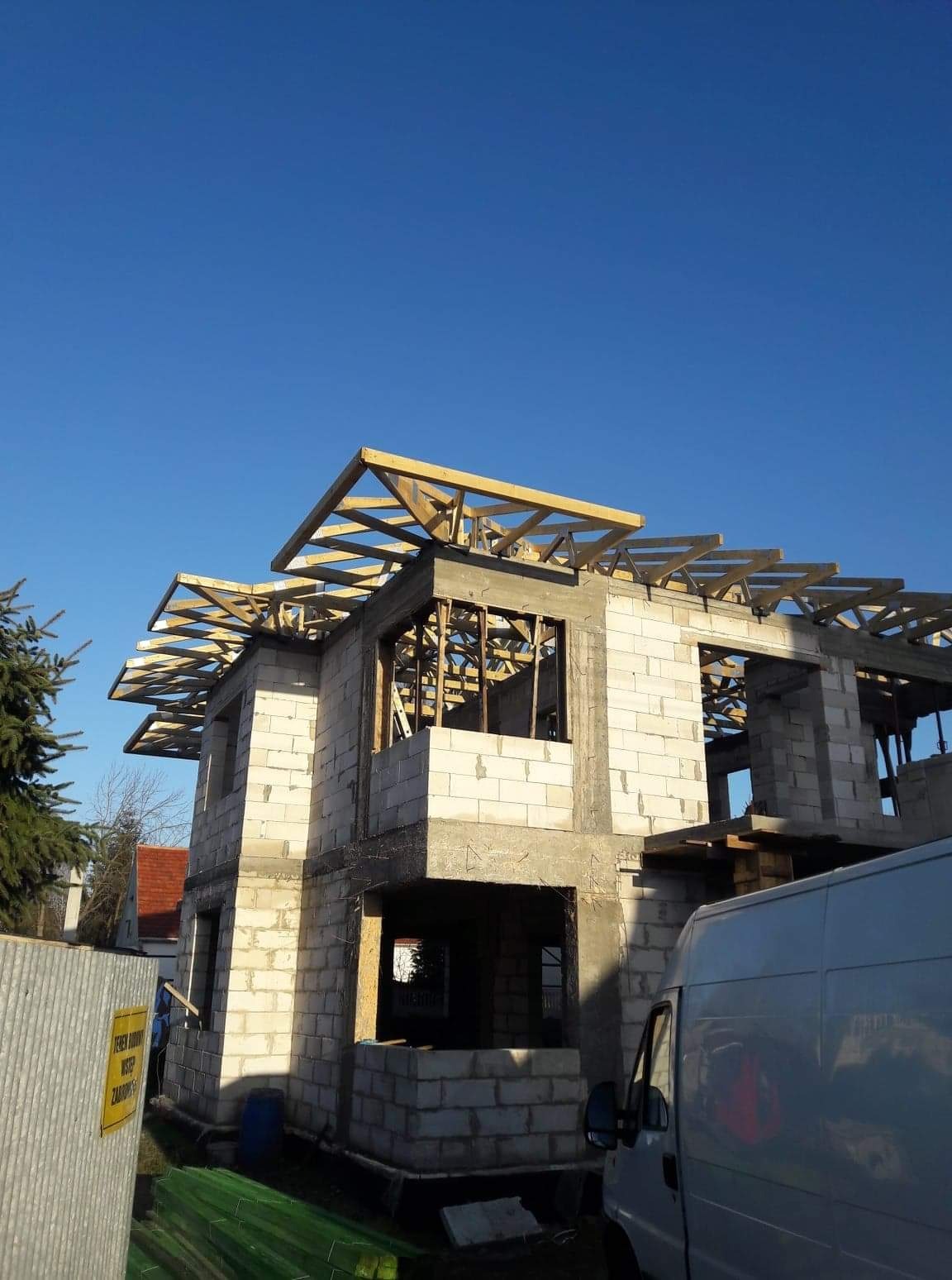 Budowa domu murowanego w Rostarzewie w trakcie montażu więźby dachowej, widoczne bloczki betonowe, drewniana konstrukcja dachu i fragment białego samochodu dostawczego.