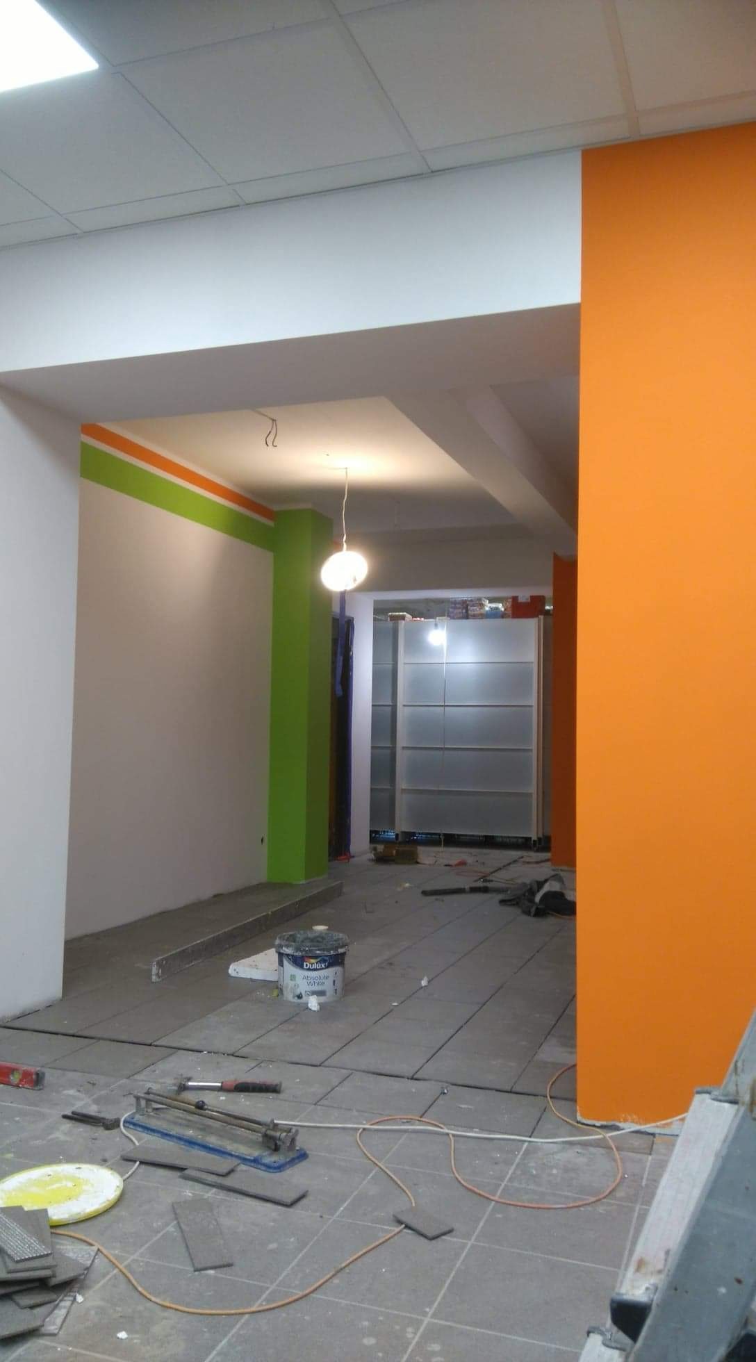 Układanie szarych płytek podłogowych w pomieszczeniu z pomalowanymi na pomarańczowo i zielono ścianami, widoczny sprzęt do cięcia płytek, puszka farby Dulux Absolute White.