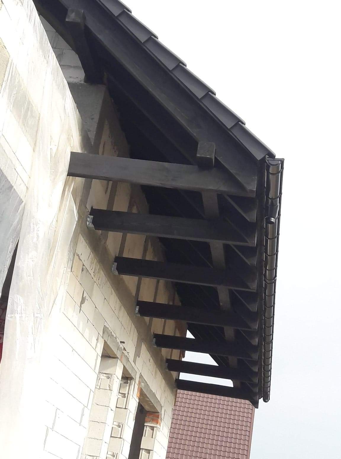 Surowy stan budowy domu z widoczną konstrukcją dachu: ciemne belki więźby dachowej wystające spod okapu z czarną blachodachówką i rynnami, ściany z białego bloczka.