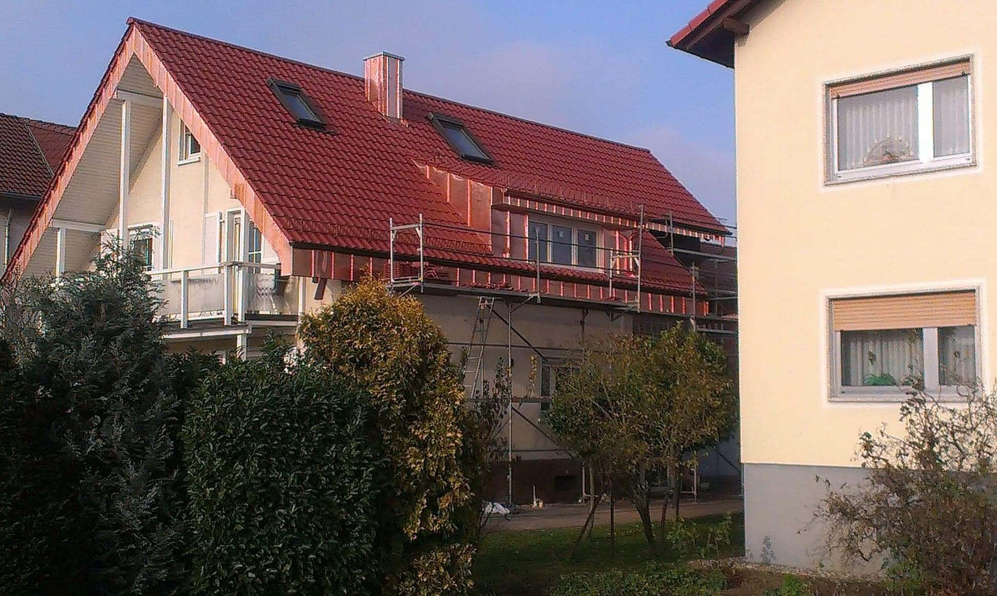 Dom jednorodzinny z czerwoną dachówką, widoczne rusztowania i elementy obróbek blacharskich w kolorze miedzi, komin również w obróbce blacharskiej, okna dachowe i balkon.