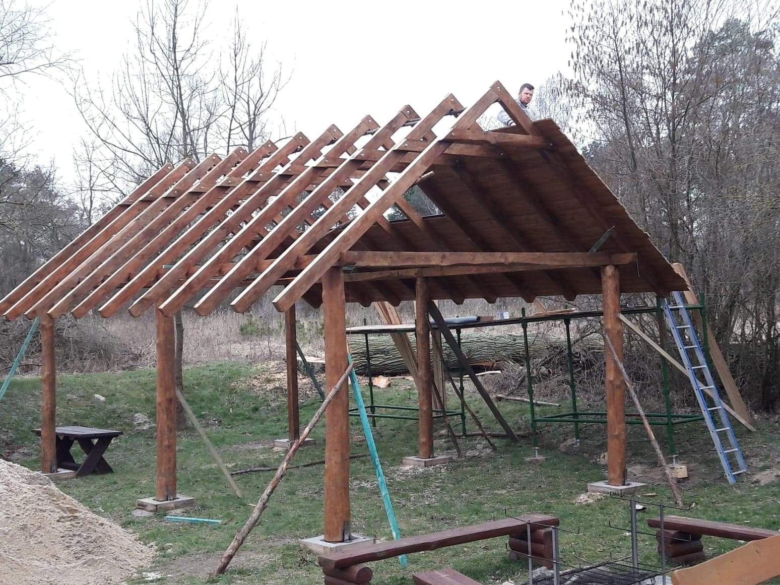 Montaż drewnianej konstrukcji zadaszenia altany, widoczne krokwie i deski poszycia, w tle las i drabina oparta o konstrukcję, na górze konstrukcji stoi pracownik.