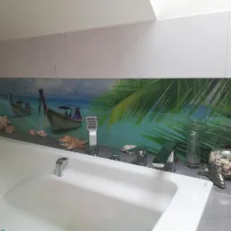 Biała wanna z baterią i słuchawką prysznicową na tle ściany z motywem tropikalnej plaży z łodziami i palmami, ozdobiona muszlami i szklanym wazonem.