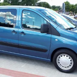 Niebieski bus osobowy zaparkowany na kostce brukowej, z widocznym odbiciem chmur na szybach.