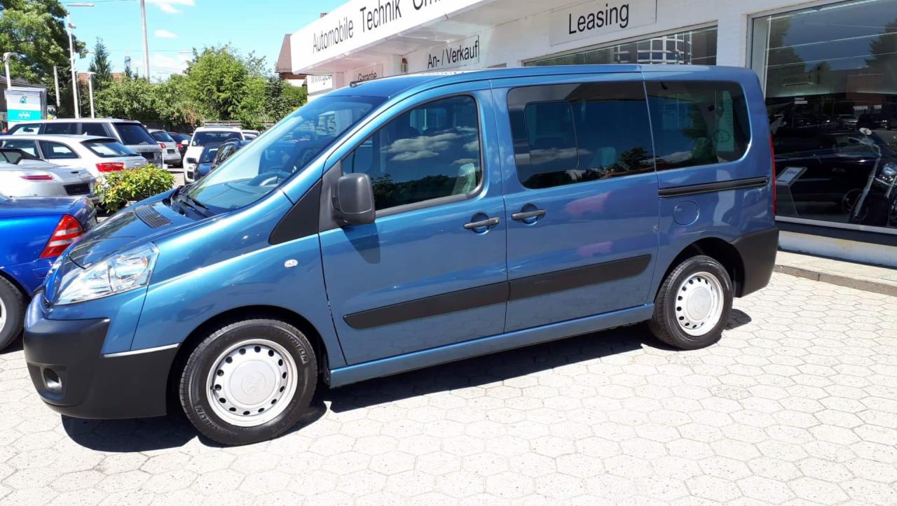 Niebieski van z przesuwnymi drzwiami, zaparkowany przed salonem samochodowym z napisem Leasing, widoczne odbicie chmur w szybach.