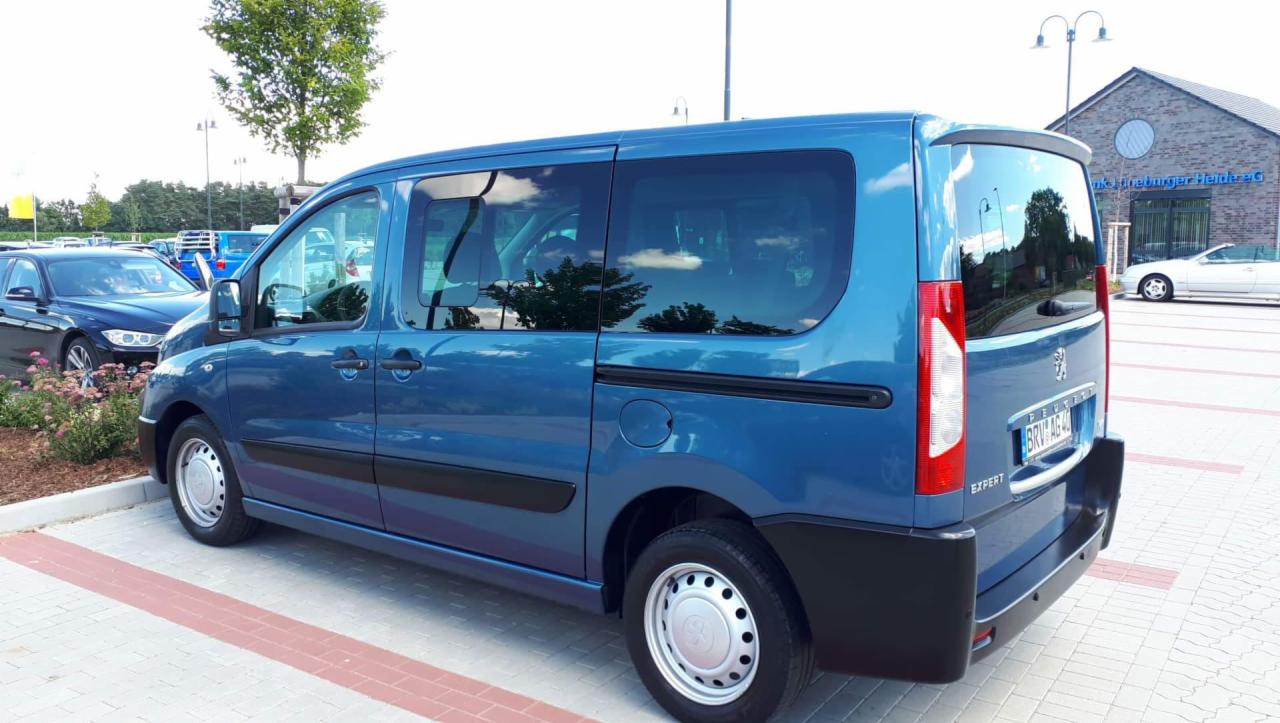 Niebieski bus Peugeot Expert zaparkowany na placu z kostki, widok z tyłu i boku, w szybach odbija się niebo i drzewa.