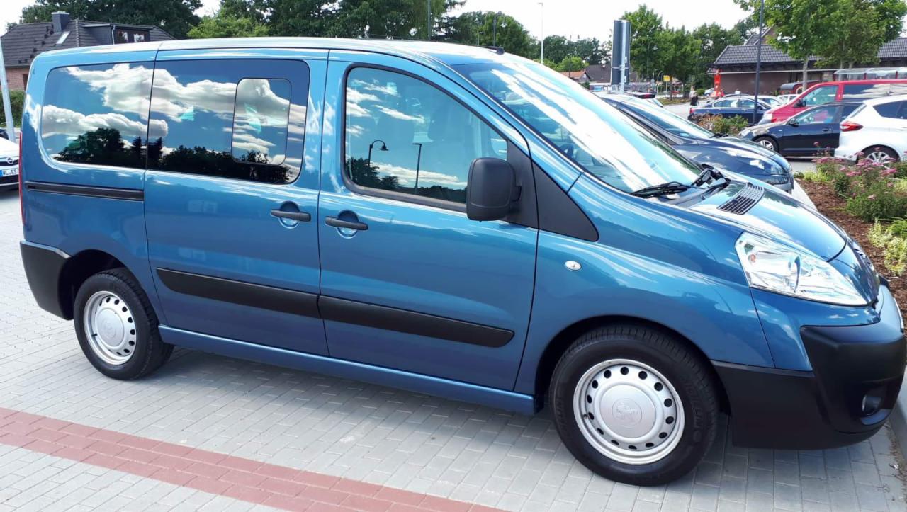 Niebieski bus Peugeot Expert Tepee z srebrnymi felgami, zaparkowany na kostce brukowej z widocznym odbiciem chmur w oknach.