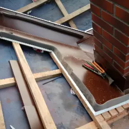 Montaż brązowych elementów blaszanych wokół komina na konstrukcji dachu, widoczne narzędzia dekarskie.