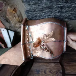 Miedziana rynna z ozdobną różą i liśćmi, widoczne spawy, na tle kamiennej ściany i drewnianej powierzchni.