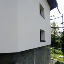 Świeżo otynkowana ściana domu z dwoma oknami, zabezpieczona folią fundamentowa, z widocznymi rusztowaniami w tle.