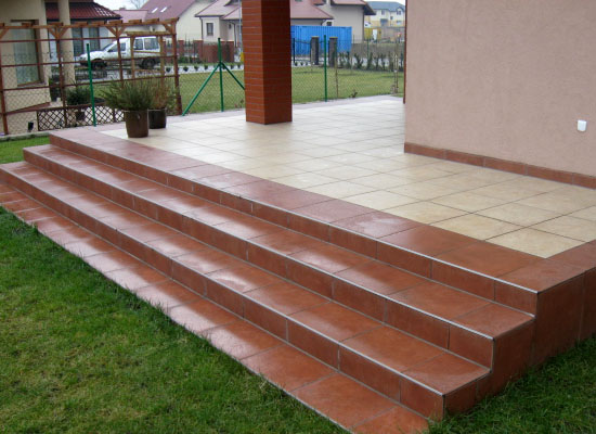 Wyłożone terakotą schody wejściowe prowadzące na taras z jasnymi płytkami, widoczne rośliny w donicach i domy w tle.