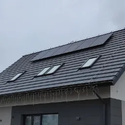 Ciemnoszary dach domu jednorodzinnego z trzema oknami dachowymi i panelami słonecznymi zamontowanymi u góry, ozdobiony lampkami świątecznymi.