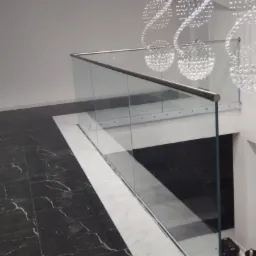 Szklana balustrada z metalową poręczą, widok z góry na marmurowe schody, dekoracyjny żyrandol w tle.