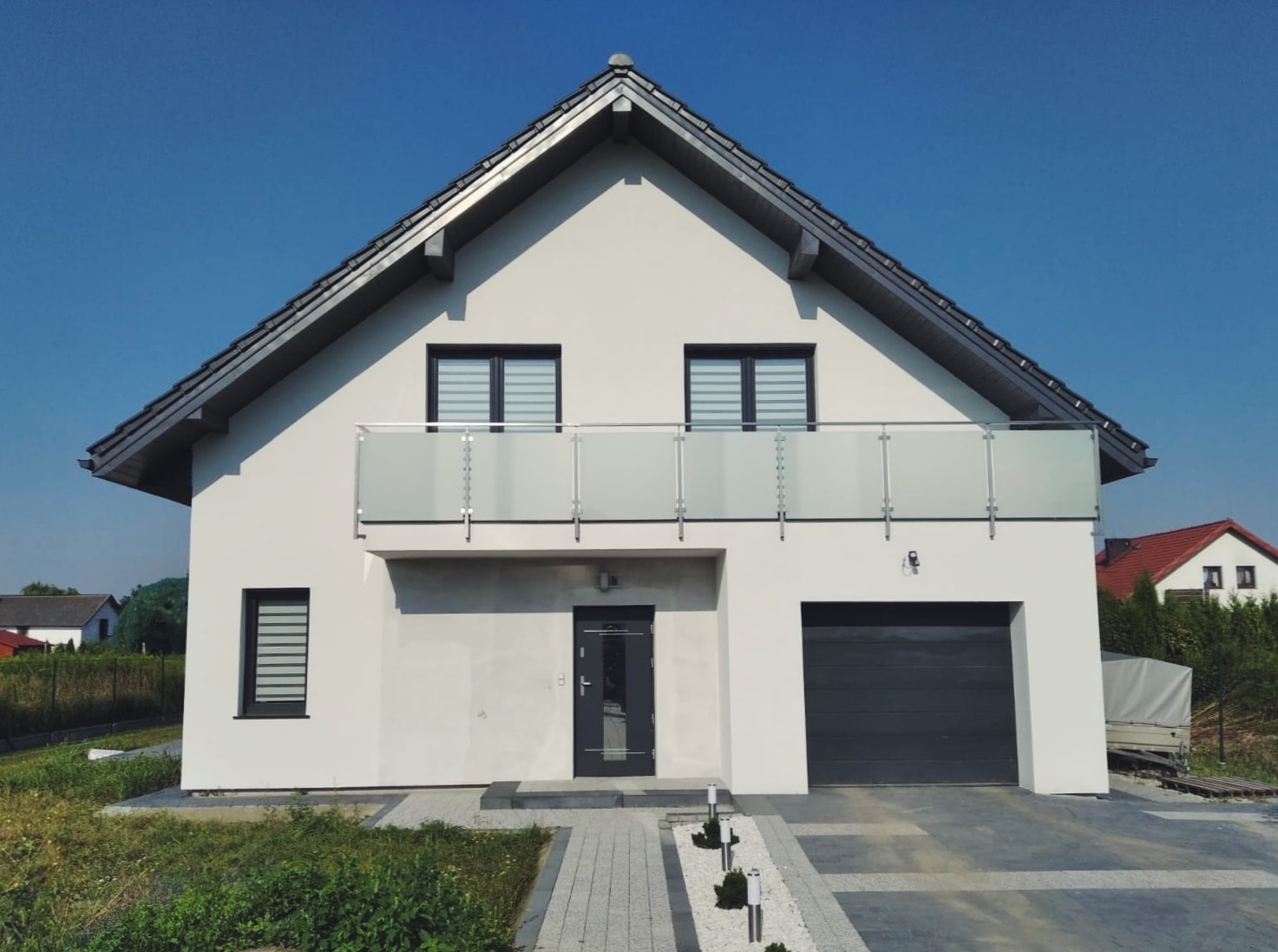 Nowoczesny dom jednorodzinny z balkonem zabezpieczonym balustradą szklaną z metalowymi słupkami, widok z przodu na tle błękitnego nieba.