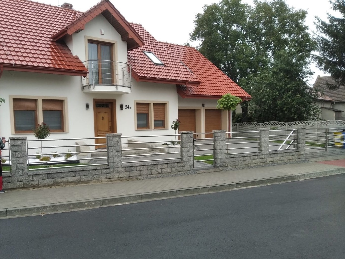 Nowoczesna posesja z białym domem i ogrodzeniem z elementami nierdzewnymi oraz kamiennymi słupkami, widok z ulicy.