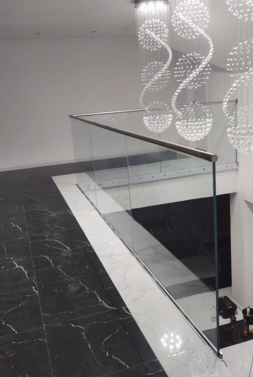 Szklana balustrada z metalową poręczą, widok z góry na marmurowe schody, dekoracyjny żyrandol w tle.