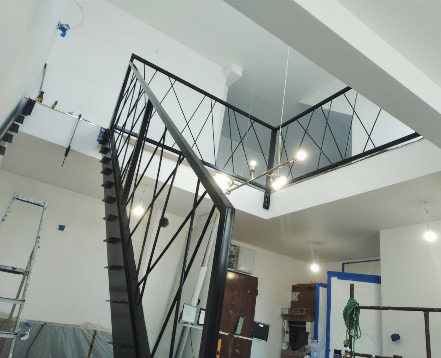 Metalowe schody z czarną, geometryczną balustradą i poręczą, widok od dołu, w tle surowe wnętrze w trakcie remontu z widoczną drabiną i zabezpieczeniami.