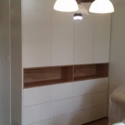 Biała, wysoka szafa z drewnianymi wnękami, oświetlona przez lampę wiszącą z trzema kloszami, na tle parkietu i krzesła z folią ochronną.