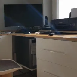 Kącik biurowy z blatem w kolorze drewna, białymi szafkami i nowoczesnym krzesłem, z komputerem stacjonarnym umieszczonym pod blatem.