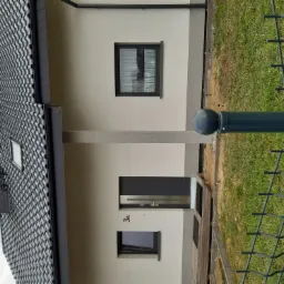 Nowoczesny dom jednorodzinny z jasną elewacją, grafitową stolarką okienną i drzwiami wejściowymi, widok zza ogrodzenia.