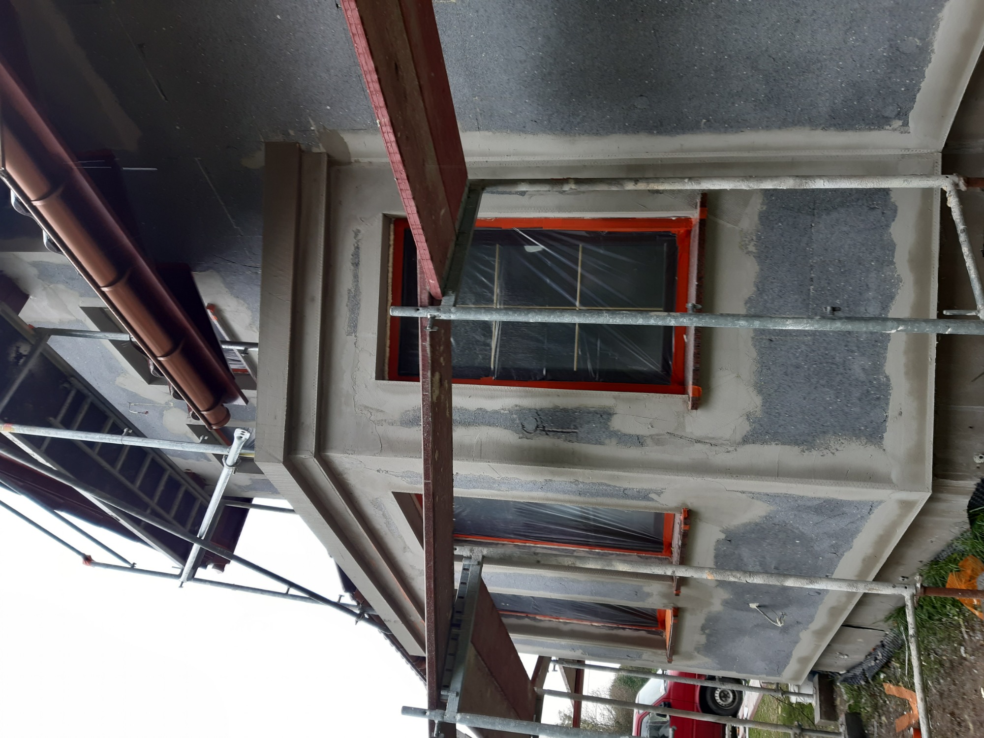 Elewacja budynku w trakcie prac: widoczne rusztowania, okna zabezpieczone folią, fragment ocieplenia i częściowo nałożony tynk w kolorze szarym, obróbka okien w kolorze pomarańczowym, rynna...