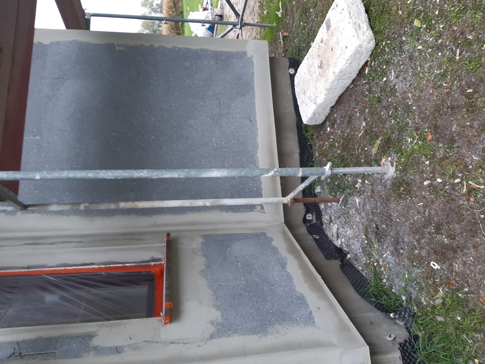 Ściana budynku w trakcie prac, widoczne rusztowanie, okno zabezpieczone folią i fragment izolacji przy gruncie.