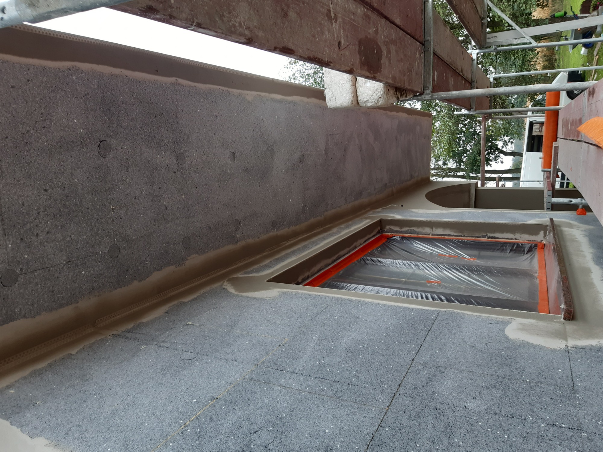 Przygotowanie do ocieplenia balkonu z użyciem szarego styropianu, zabezpieczonego folią, widoczne rusztowanie i fragment ściany elewacyjnej z nałożonym tynkiem.