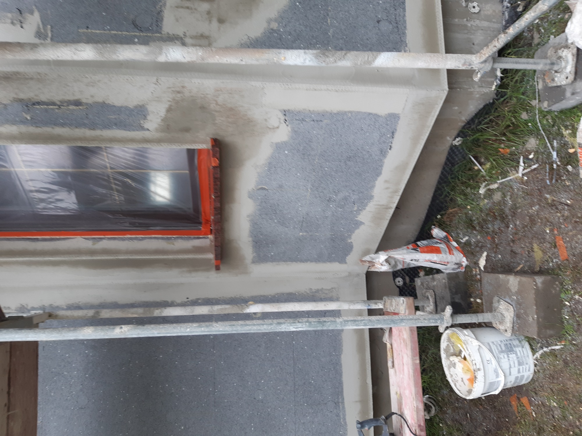 Fragment elewacji w trakcie prac, widoczne rusztowanie, okno zabezpieczone folią, świeżo nałożona warstwa izolacyjna o szarej fakturze, częściowo jeszcze nieotynkowana.