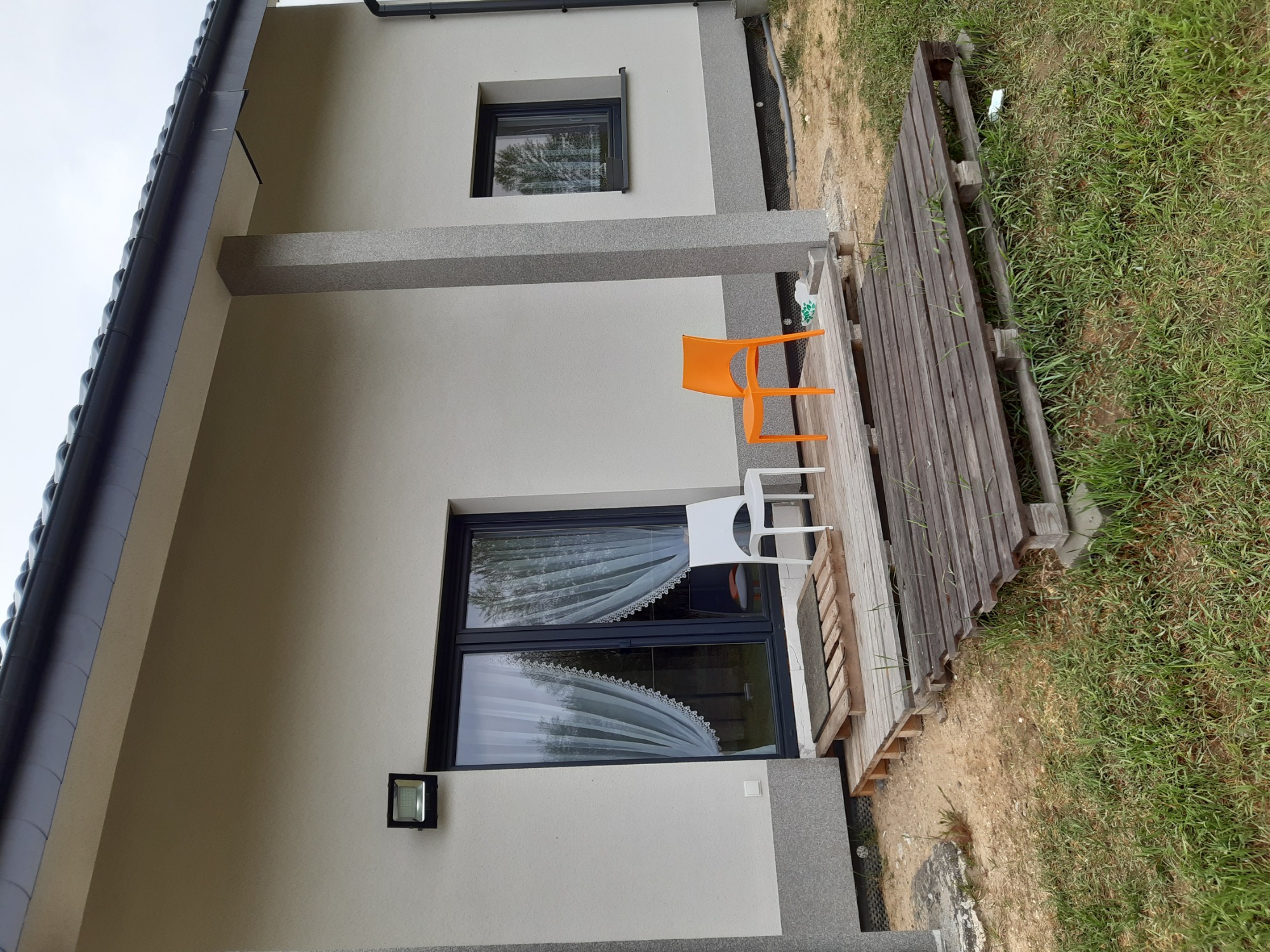 Szara elewacja domu jednorodzinnego z dwoma oknami, na zewnątrz dwa krzesła (białe i pomarańczowe) stojące na drewnianym podeście.