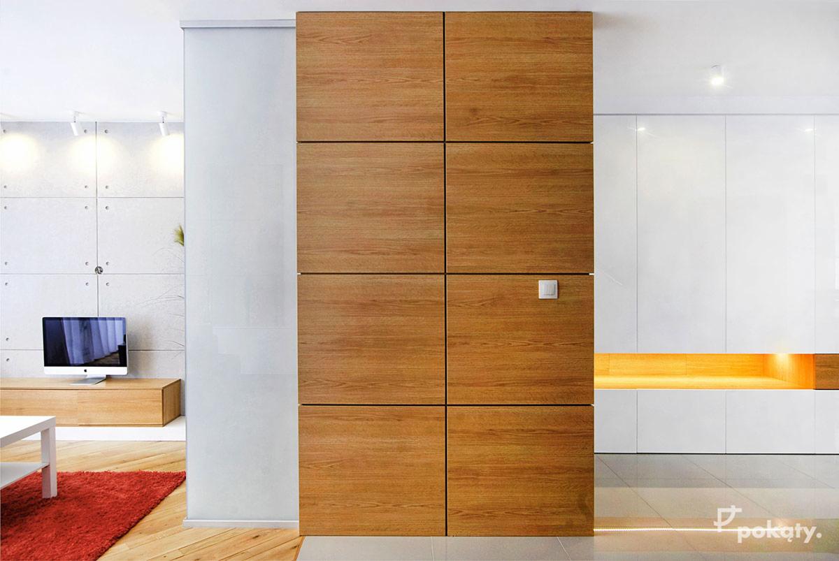 Nowoczesny salon z drewnianą ścianą działową z kwadratowych paneli, białą ścianą z wnęką podświetloną ciepłym światłem, betonową ścianą z telewizorem i czerwonym dywanem.