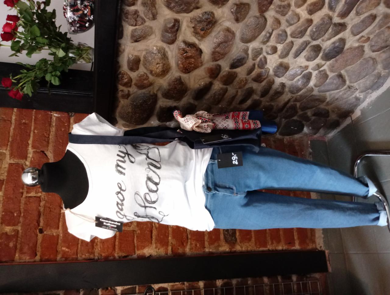 Manekin prezentuje białą koszulkę z napisem 'gave my heart' oraz jasnoniebieskie jeansy, całość na tle ściany z kamienia i cegły.