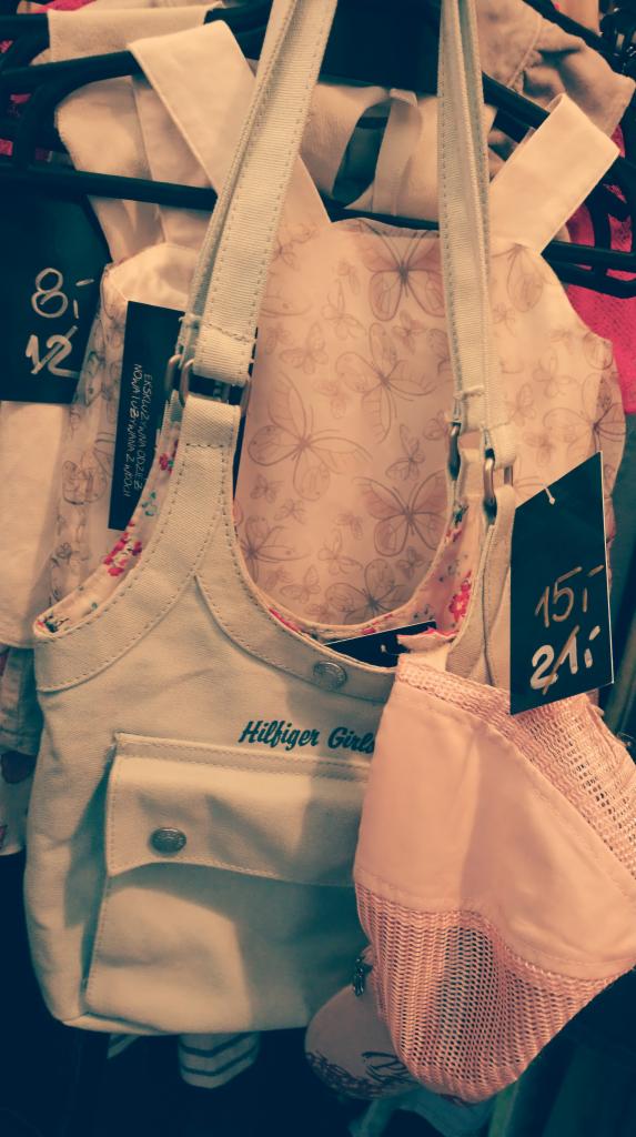 Jasna sukienka z motywem motyli, dżinsowa torba na ramię z napisem Hilfiger Girls i różowa czapka z daszkiem, wiszące na wieszaku w sklepie.