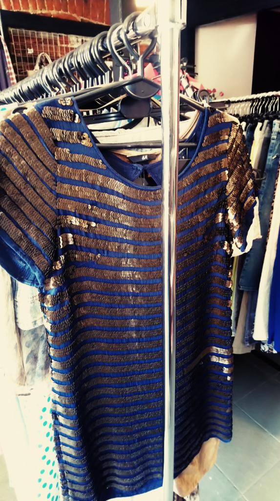 Niebieska bluzka w złote cekiny na wieszaku, widoczne inne ubrania w tle.