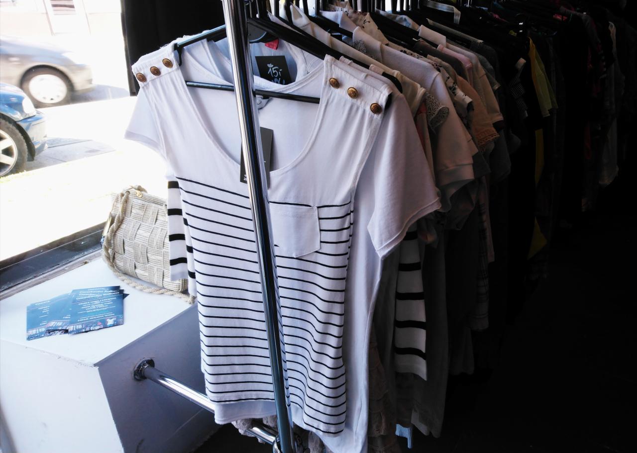 Rząd ubrań na wieszakach, z przodu biała koszulka w czarne paski z kieszonką i ozdobnymi guzikami na ramionach, obok tkana torebka i ulotki.