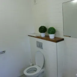 Nowoczesna łazienka z białymi płytkami imitującymi cegłę, podwieszana toaleta z ukrytą spłuczką, drewniana półka z dwoma małymi sztucznymi roślinami, szara podłoga z płytek.