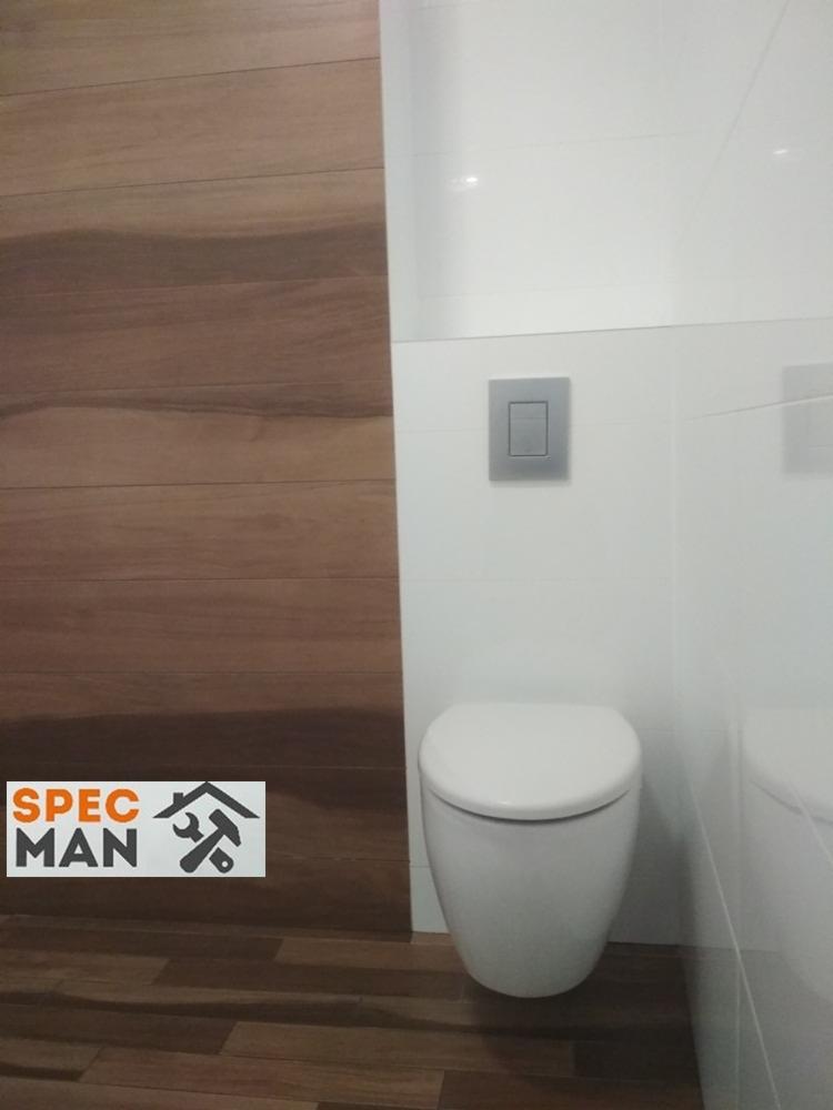 Nowoczesna toaleta wisząca w łazience z drewnopodobną okładziną ścienną po lewej stronie i białymi płytkami po prawej, widoczny szary przycisk spłuczki.