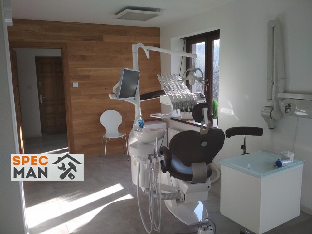Nowoczesny gabinet stomatologiczny z drewnianą okładziną ścienną, wyposażony w biały fotel dentystyczny, unit z licznymi narzędziami i monitor oraz szafkę pomocniczą z przezroczystym blatem.